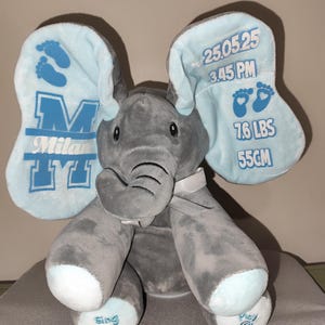 May include: A gray plush elephant toy with light blue accents on the ears and feet. The elephant's ears display the name "Milan", footprints, and birth details: 7.6 lbs and 55cm. The toy has "Sing" and "Play" buttons.