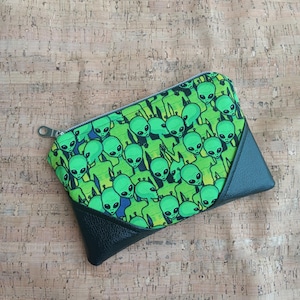 May include: A rectangular zippered pouch featuring a repeating pattern of green alien figures on a lime green background. The pouch has a black leather-like corner and a silver zipper.
