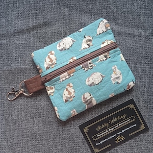 May include: A teal zippered pouch featuring a repeating pattern of cartoon bison. The pouch has a brown zipper and a brown wrist strap with a silver clasp. A business card with the text "Stitchy Witchings" is in the bottom right corner.