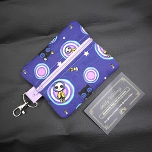 May include: A purple zippered pouch with a cartoon character and cat pattern. The pouch has a lavender zipper and a silver clasp. A business card with the text "Stitchy Witchings" is next to the pouch.