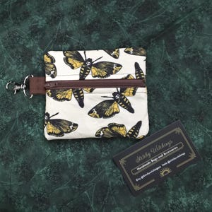 May include: A small, square coin purse with a white background and a repeating pattern of black and yellow moth illustrations. The purse has a brown zipper and a silver clasp. A business card with the text "Stitchy Witchings" is also visible.