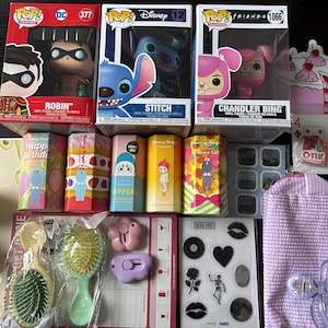 Puede incluir: Una colección de varios artículos que incluyen figuras Funko Pop de Robin, Stitch y Chandler Bing. También incluye cepillos para el cabello, pegatinas y pequeñas cajas de regalo con personajes de dibujos animados. También se ve una bolsa morada y naipes.