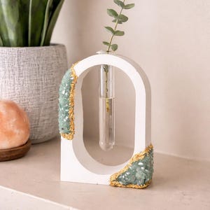 Puede incluir: Jarrón blanco en forma de arco con un tubo de vidrio transparente para un solo tallo. El jarrón está adornado con racimos de cristal verde claro y ribetes dorados. Una ramita de vegetación está en el tubo.