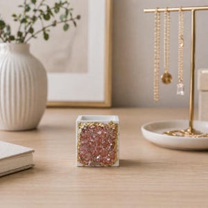 May include: A small, square white ceramic container with a pink crystal and gold leaf design. The container is on a wooden surface, with a vase, framed artwork, and jewelry stand in the background. The jewelry stand holds several gold necklaces.
