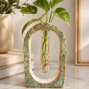 May include: An oval-shaped propagation station featuring a white frame with textured light green crystal-like accents and gold leaf detailing. A clear glass test tube is suspended in the centre, holding a green plant with variegated leaves.