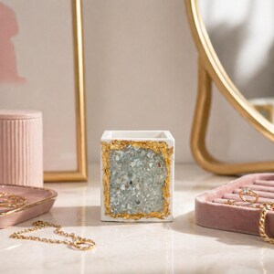 May include: A white square decorative container with a gold leaf border and a center filled with blue-gray crushed stone. The container is on a white surface with other decorative items, including a pink jewelry tray and a gold-framed mirror.
