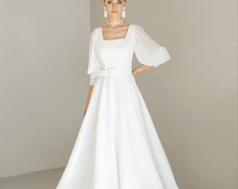 May include: A white wedding dress with a square neckline and a flowing skirt. The dress features sheer sleeves and a belted waist. The model is wearing pearl earrings and a necklace. The dress is elegant and classic.