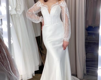 May include: A white wedding dress with a fitted bodice and a mermaid silhouette. The dress features a plunging neckline and sheer, long sleeves with intricate lace detailing. The bride is wearing a silver necklace.