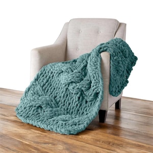 Puede incluir: Una manta de punto grueso de color turquesa sobre un sillón beige. La manta tiene un patrón tejido y un aspecto suave y texturizado. La silla está sobre un suelo de madera.