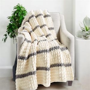 Pode incluir: Uma manta creme texturizada com listras cinza horizontais, drapeada sobre uma poltrona cinza claro. A manta tem uma aparência espessa e aconchegante, sugerindo uma textura macia e quente. O fundo inclui vegetação e uma mesa lateral branca.