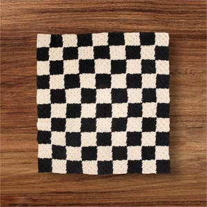 May include: A square, black and cream-colored checkerboard pattern rug. The rug is made of a textured material and is placed on a wooden surface. The design features alternating black and cream squares.