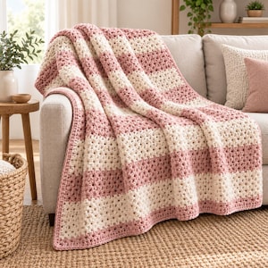 May include: A crochet blanket featuring alternating horizontal stripes of dusty rose pink and cream white yarn. The textile has a textured, open-weave stitch pattern throughout, creating a soft, layered appearance as it drapes over a piece of furniture.