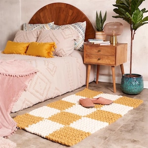 Può includere: Un tappeto fatto a mano con un motivo a scacchiera bianco e giallo senape è appoggiato su un pavimento grigio. Il tappeto è realizzato con filato spesso e grosso. Delle pantofole rosa sono appoggiate sul tappeto vicino a un letto con lenzuola rosa e cuscini gialli. Sono visibili anche un comodino in legno e una pianta in vaso.