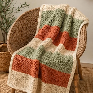 May include: A rectangular crochet blanket featuring horizontal stripes in cream, sage green, and burnt orange. The textile has a textured, bumpy stitch pattern throughout and is finished with a simple cream-coloured border along all four edges.