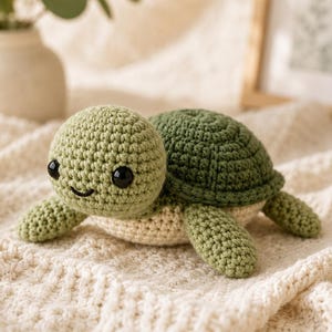 May include: A small, handmade crochet turtle toy featuring a light sage green head and flippers, a darker forest green shell, and a cream-coloured underside. The turtle has two round, black plastic safety eyes and a stitched black smile.