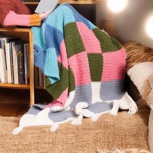 Può includere: Una colorata coperta all'uncinetto fatta a mano con un design patchwork. La coperta presenta quadrati rosa, blu, verdi, grigi e bianchi. La coperta ha un bordo con frange ed è drappeggiata su una superficie.