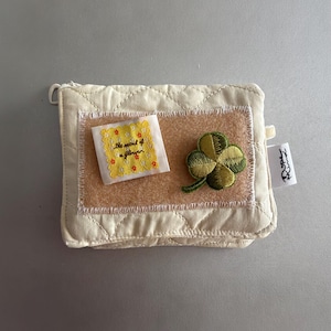 Puede incluir: Un estuche acolchado de color crema con un parche rectangular de tela beige. Sobre el parche hay una pequeña etiqueta cuadrada blanca con motivos florales amarillos y el texto "the scent of a flower", junto a un bordado de trébol de cuatro hojas.