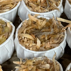 May include: Close-up of several white paper cups filled with dried, light brown, and tan pieces. The cups are arranged closely together, showcasing the texture and color variations of the contents. The image is well-lit, highlighting the details of the dried product.