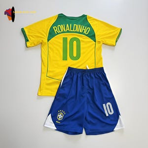 Puede incluir: Un conjunto de camiseta de fútbol amarilla y verde y pantalones cortos azules. La camiseta tiene el nombre "RONALDINHO" y el número "10" en la espalda. Los pantalones cortos también tienen el número "10". El conjunto incluye el logo de la bandera brasileña.