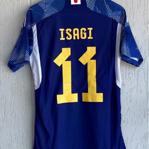 Può includere: Una maglia da calcio blu e bianca con il nome "ISAGI" e il numero "11" in giallo. La maglia ha un'emblema della bandiera giapponese sulla parte superiore della schiena ed è appesa a una gruccia bianca.