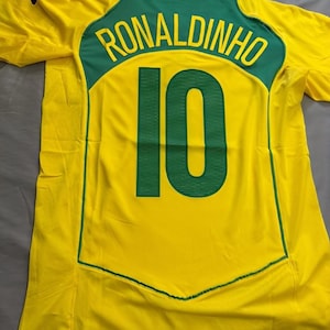 May include: A yellow football shirt with green detailing, displaying the name "RONALDINHO" and the number "10" in green. The shirt has a green collar and trim. The Fandom Supply Shop logo is visible in the top left corner.
