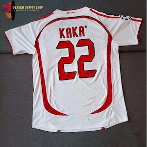 Puede incluir: Camiseta de fútbol blanca con detalles rojos, con el nombre "KAKA'" y el número "22" en rojo. La camiseta tiene cuello redondo y mangas cortas. La camiseta tiene un logo en la esquina superior izquierda.
