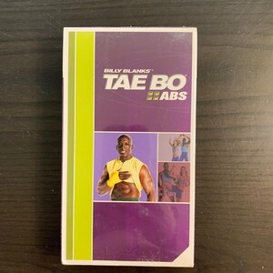 May include: A rectangular VHS tape with the title "TAE BO ABS" in white and yellow lettering. The tape features a purple background with a photo of a man in a yellow shirt. The tape has a green vertical stripe on the left side.
