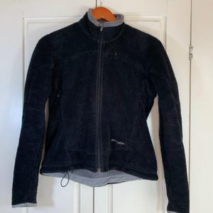 May include: Black fleece jacket with a full-length zipper, a gray collar, and a small logo on the chest. The jacket has a drawstring at the bottom hem and is hanging on a wooden hanger against a white door.