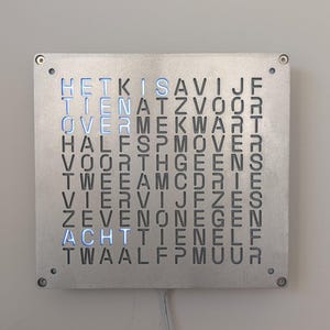 Pode incluir: Um relógio de parede quadrado em metal escovado com letras iluminadas em azul. O mostrador mostra as horas em holandês. O relógio está montado numa parede cinza claro. As dimensões não são visíveis.