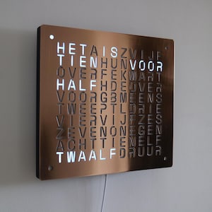 Pode incluir: Um relógio de parede quadrado com acabamento em cobre metálico, apresentando uma grade de letras holandesas. O relógio mostra as horas iluminando letras específicas em branco, exibindo a frase HET IS TIEN VOOR HALF TWAALF.