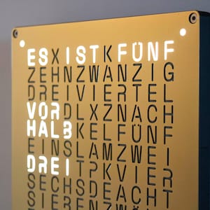 Peut inclure: Une horloge murale rectangulaire avec une façade métallique dorée présentant une grille de lettres noires en allemand. Les mots ES, IST, VOR, HALB et DREI sont éclairés par une lumière blanche vive, indiquant l'heure sur la grille.