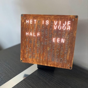 Può includere: Un orologio quadrato in metallo marrone arrugginito con le parole "HET IS VIJF VOOR HALF EEN" illuminate in bianco. L'orologio è montato su una base nera e poggia su una superficie scura.