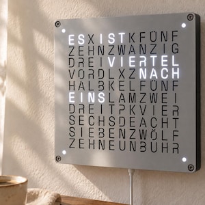 Pode incluir: Um relógio de parede quadrado com uma face prateada apresentando uma grade de letras alemãs pretas. O relógio exibe a hora usando letras brancas iluminadas que formam VIERTEL NACH EINS. Quatro pequenas luzes brancas estão posicionadas nos cantos.