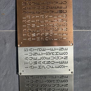 Könnte beinhalten: Drei Metall-Wanduhren in Kupfer, Silber und Dunkelgrau. Jede Uhr hat eine rechteckige Form mit abgerundeten Ecken und die Zeit in vertikalen Textzeilen eingraviert. Die Uhren sind mit kleinen Löchern montiert.