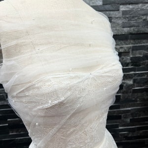 May include: White lace fabric with a sheer overlay and pearl embellishments. The fabric is draped over a dress form.