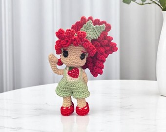 May include: A handmade crochet doll with red hair, green overalls, and red shoes. The doll has a strawberry design on the front of the overalls and a green leaf-shaped bow. The doll is approximately 6 inches tall.