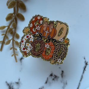 May include: A decorative pin with the phrase "God is Good" in a mix of fonts and patterns. The pin features a pumpkin with a cross, floral and leopard print designs, and gold glitter accents. The color palette includes orange, brown, white, and gold.