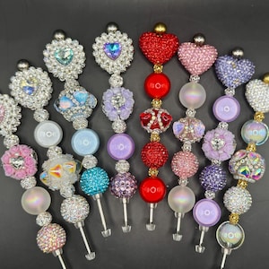 May include: A collection of decorative bottle stoppers with various designs. Each stopper features a decorative top with rhinestones, beads, and heart or floral shapes. The color palette includes pink, red, blue, and purple, with silver and gold accents.