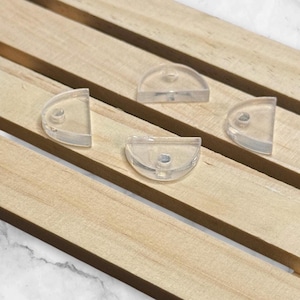 May include: Four clear, semi-circular plastic clips with a small hole in the center, arranged on a light brown wooden surface. The clips are translucent, allowing the wooden texture to be partially visible through them.
