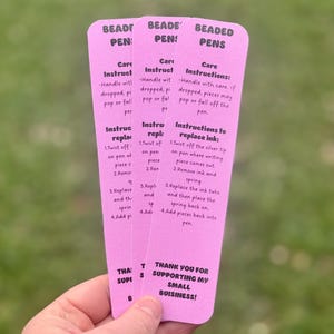 May include: Three pink instruction cards for beaded pens are held in a hand. The cards have text that includes care instructions and instructions for replacing ink. The bottom of the card says, "Thank you for supporting my small business!"