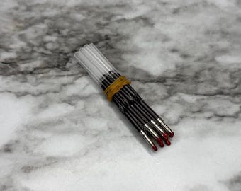 May include: A bundle of black ballpoint pen refills with red ink tips, held together by a yellow rubber band. The clear plastic tubes of the refills are visible. The background is a marbled gray and white surface.