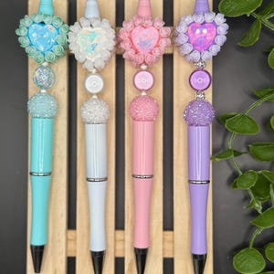 May include: Four decorative pens in pastel colors: turquoise, light blue, pink, and purple. Each pen features a heart-shaped embellishment surrounded by roses, with a sparkling bead and a decorative top. The pens are displayed on a wooden stand.
