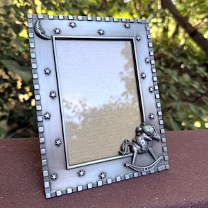 May include: A rectangular silver-toned metal picture frame featuring a brushed finish. The border is decorated with embossed stars and a crescent moon. A three-dimensional rocking horse figure is attached to the bottom right corner of the frame.