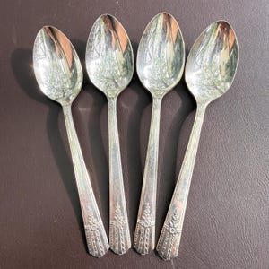 May include: Four silver spoons with oval bowls and long, decorated handles. The spoons reflect the surrounding environment. The handles have a floral design at the base. The spoons are arranged on a brown surface.