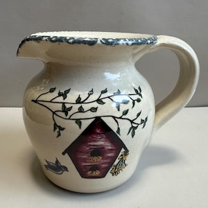 May include: Cream-colored ceramic pitcher with a decorative birdhouse design. The pitcher features a dark red birdhouse, green leaves, and a small blue bird. The rim has a blue and white pattern, and the handle is curved.