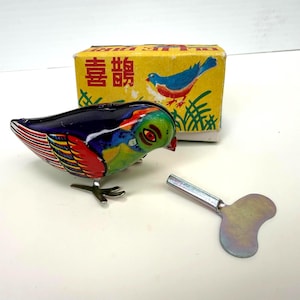 May include: A colorful tin wind-up bird toy with blue, red, and green patterns, featuring a metal winding key and a yellow cardboard box with a blue bird illustration and the Chinese characters 喜鵲 printed on the front.