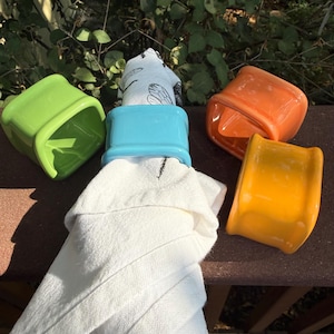 May include: A set of four square ceramic napkin rings in bright lime green, light blue, orange, and yellow. One light blue napkin ring holds a white cloth napkin featuring a black line drawing of a dragonfly. The rings have a smooth, glossy finish.