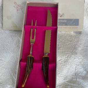 May include: A vintage carving set in a box. The set includes a carving fork and knife with serrated blades. The handles have a brown and yellow pattern. The box is white and pink with the text "Sheffield Cutlery Set".