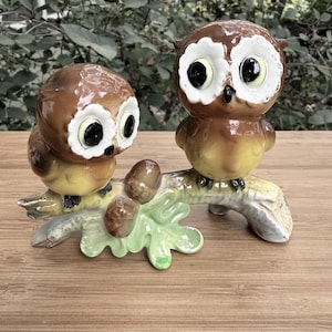 May include: A ceramic figurine featuring two brown owls with large white-rimmed eyes perched on a textured branch. The branch includes a light green oak leaf and two brown acorns. The owls have yellow-toned bellies and dark brown wings.