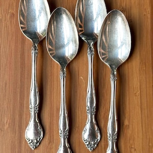 May include: Four silver-toned metal spoons arranged vertically. Each spoon features an oval bowl with a reflective surface and a decorative handle with embossed floral details at the base and along the stem, showing a consistent metallic finish.
